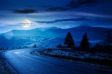 Gece dağlarda köy yolu. Sonbaharın sonlarında güzel bir kır manzarası. Yol kenarındaki köknar ağaçları dolunay ışığında. Manzaralı mavi gökyüzü. Kardaki uzak tepenin üzerindeki bulutlar.