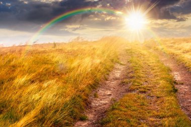 Yazın gün batımında sisli arazide toprak yol manzarası. Akşam ışığında bulutlu gökyüzünün altındaki tepelerde sarı çimenler. gizemli atmosfer kırsal kompozit fotoğraf