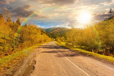 Sonbaharda gün batımında dağ manzarası boyunca yol. Sonbahar renklerinde orman mavi gökyüzünün altında ve akşam ışığında gökkuşağı. Uzakta karla kaplı zirve. Kırsal Vadi manzarası