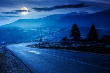 Gece dağlarda köy yolu. Sonbaharın sonlarında güzel bir kır manzarası. Yol kenarındaki köknar ağaçları dolunay ışığında. Manzaralı mavi gökyüzü. Kardaki uzak tepenin üzerindeki bulutlar.