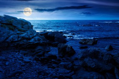 weekend on a rocky beach at the black sea under open dark sky at night. beautiful coastline in summer. seascape scenery with clouds above horizon in full moon light. mysterious weather