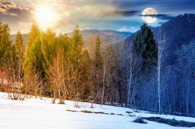 Karla kaplı yamaçta karışık ormanlarla kaplı dağ manzarası. Gündüz ve gece zaman kavramını değiştirir. Bulutlu gökyüzü, ilkbahar ekinoksunda güneş ve ay. ikilik ve geçiş şablonu. çıplak ağaçlar manzarası