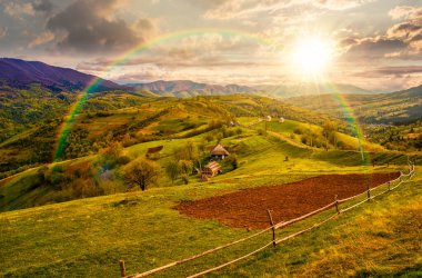 Gün batımında kırsal bölgedeki yamaçta ekilebilir ve ahşap çitlerin manzarası. İlkbaharda akşam ışığında güzel dağ manzarası. Gökkuşağının altında hikaye anlatımı sinemasal görüntü