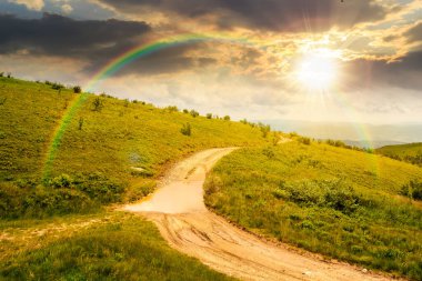 Yaz günbatımında dağların arasından geçen toprak yol. Mavi gökyüzünün altındaki yeşil yuvarlanan tepelerin manzarası ve akşam ışığında bulutlar. Gökkuşağının altında kurumsal bir duvar kağıdı var.