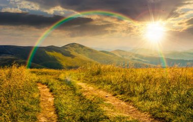 Gün batımında tepedeki çimenli çayırların arasından toprak yol. Akşam ışığında bulutlu gökyüzünün altındaki yeşil dağlarda güzel bir yaz manzarası. gökkuşağıyla kaplı sonsuz nostalji kitabı.