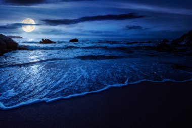 Güzel boş kumlu sahil geceleri denizde. Dolunay ışığında kayanın yanındaki kıyıya doğru yuvarlanan beyaz köpük. Mükemmel yaz tatili konsepti. Harika, gizemli hafta sonu fantezisi. düşük açı