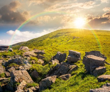 Yazın gün batımında mavi gökyüzünün altında güneşli dağ manzarası. Akşam ışığında, dik yamaçta yemyeşil çimenler ve keskin kayalar. Gökkuşağının altındaki dağlık alanda güzel bir manzara