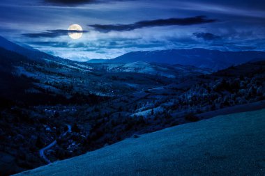 Baharda geceleri dağlık kırsal alan. Dolunay ışığında vadideki köy. Yuvarlanan tepelerdeki çimenli kırsal alanlar. Kar lekeleri olan uzak bir sırt. gizemli bulutlu hava
