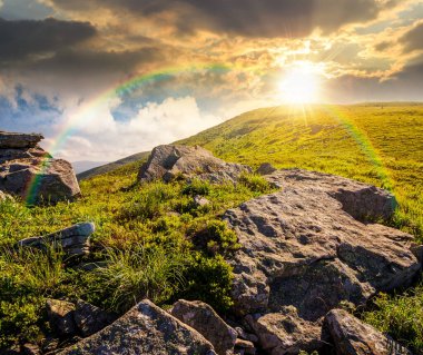 Yazın gün batımında mavi gökyüzünün altında güneşli dağ manzarası. Akşam ışığında, dik yamaçta yemyeşil çimenler ve keskin kayalar. Gökkuşağının altındaki dağlık alanda güzel bir manzara