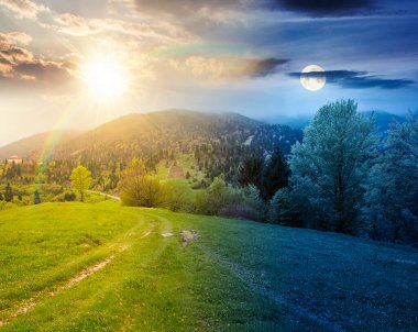 Dağ manzarasının kırsal kesimi. Gündüz ve gece değişiyor. Baharda güneşli ve aylı sisli bir hava. Bir tepedeki yemyeşil otlaktan geçen toprak yol. Yaprak döken ağaçları olan alp manzarası