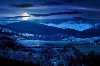 autumn landscape of rural region in mountains at night. rolling hills with green alps in dappled full moon light. serene countryside under cloudy sky. finance investment or abstract concepts
