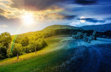 Dağ manzarasında çimenli kırsal alanlar. Gündüz ve gece. Güneş ve ay kırsal yoldan ormana yuvarlanan tepelerin arasından geçiyor. Yaz gündönümünde gökyüzünde bulutlar olan güzel bir manzaranın resmi.