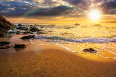 Denizde kumlu kumsalda güzel bir gün batımı. Akşamüstü gökkuşağıyla kayanın yanındaki kıyıya doğru yuvarlanan beyaz köpük. Mükemmel yaz tatili konsepti. Harika bir hafta. düşük açı görünümü
