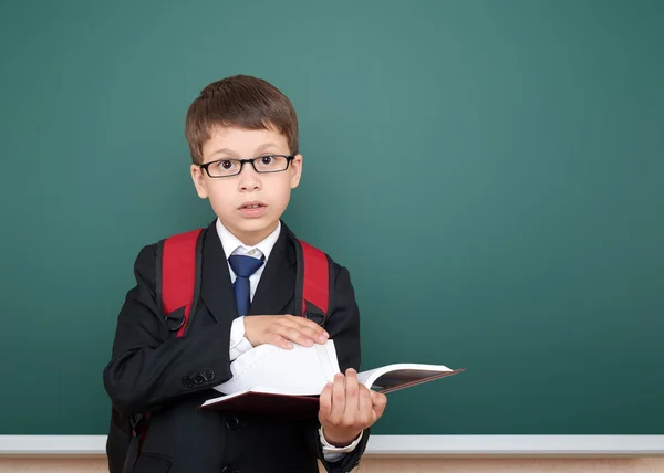 School jongen portret in zwart pak op groene schoolbord achtergrond met rode rugzak en boek, onderwijsconcept — Stockfoto