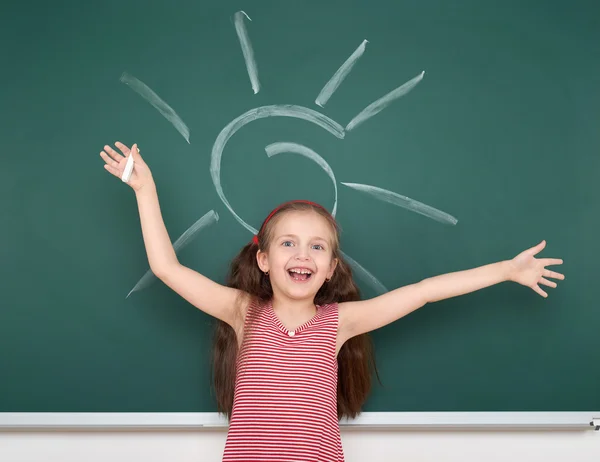 Schoolgirl child in red striped dress drawing sun on green chalkboard background, summer school vacation concept — Stock Photo, Image