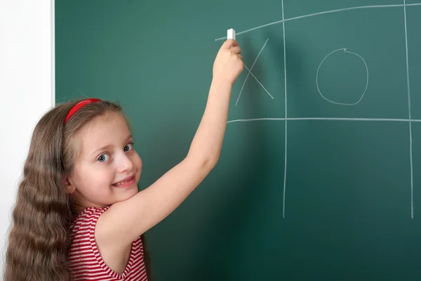 Schoolgirl child in red striped dress drawing tic tac toe on green chalkboard background, summer school vacation concept — Stock Photo, Image