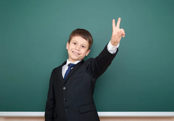 Niño de la escuela en traje negro muestran gesto de dos dedos en el fondo de pizarra verde, concepto de educación —  Fotos de Stock