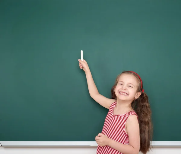 Schoolgirl child in red striped dress point and drawing on green chalkboard background, summer school vacation concept — Stock Photo, Image