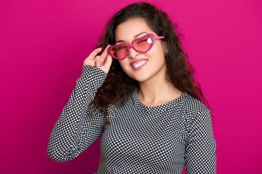 Kadın göz kırpması, güzel bir portre, pembe arka planda poz, uzun kıvırcık saçlar, kalp şeklinde güneş gözlükleri, cazibe konsepti.