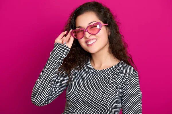 Kadın göz kırpması, güzel bir portre, pembe arka planda poz, uzun kıvırcık saçlar, kalp şeklinde güneş gözlükleri, cazibe konsepti.