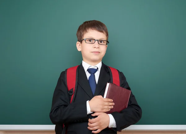 School boy portrait in black suit on green chalkboard background with red backpack and book, education concept — Stock Photo, Image