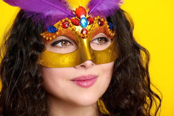 Woman face closeup in carnival masquerade mask with feather, beautiful girl portrait on yellow color background, long curly hair — Stock Photo, Image