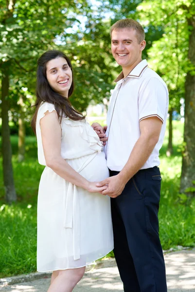Kobieta w ciąży z mężem pozowanie w parku miejskiego, portret rodziny, w sezonie letnim, Zielona trawa i drzewa — Zdjęcie stockowe
