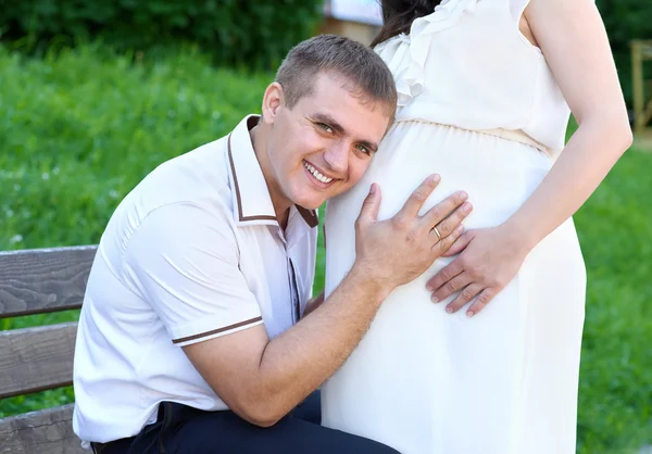 pregnant woman and husband, man listen belly and talk with baby, happy family, couple in city park, summer season, green grass and trees