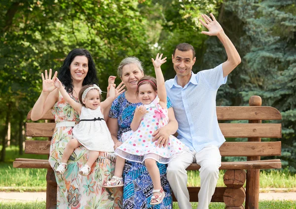 Família grande senta-se no banco de madeira no parque da cidade e acenando, estação de verão, criança, pai e avó, grupo de cinco pessoas — Fotografia de Stock