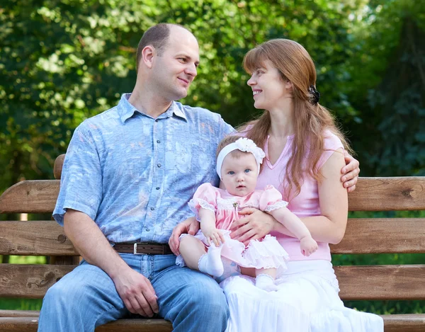 Retrato de família feliz com bebê menina ao ar livre, sente-se no banco de madeira no parque da cidade, temporada de verão, criança e pai — Fotografia de Stock