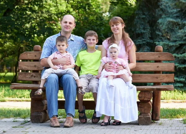 Retrato da família feliz no exterior, grupo de cinco pessoas sentam-se no banco de madeira no parque da cidade, estação do verão, criança e pai — Fotografia de Stock