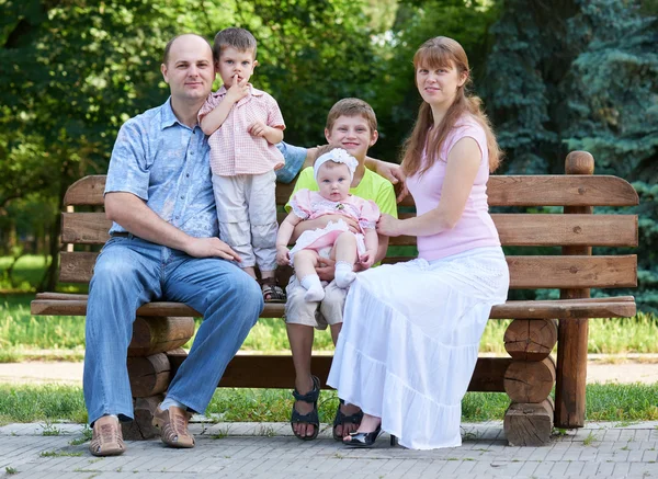 Retrato da família feliz no exterior, grupo de cinco pessoas sentam-se no banco de madeira no parque da cidade, estação do verão, criança e pai — Fotografia de Stock