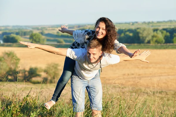 Feliz pareja con girasoles divertirse y caminar a lo largo de la carretera al aire libre, chica montando en su espalda y volar - viajes románticos, senderismo, turismo y concepto de personas — Foto de Stock