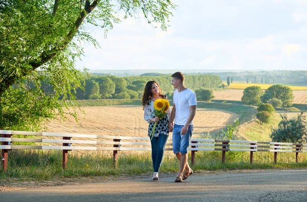 Glückliches Paar mit Sonnenblumen, die Spaß haben und auf der Landstraße im Freien spazieren gehen - romantisches Reisen, Wandern, Tourismus und Personenkonzept — Stockfoto