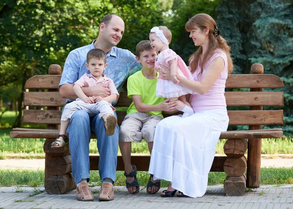 Retrato da família feliz no exterior, grupo de cinco pessoas sentam-se no banco de madeira no parque da cidade, estação do verão, criança e pai — Fotografia de Stock