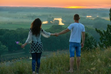 Romantik Çift tepe üzerinde günbatımında ülke açık, kız vahşi çiçek, güzel manzara ve parlak sarı gökyüzü, aşk hassasiyet kavramı, Yetişkin gençler buket ile yürümek