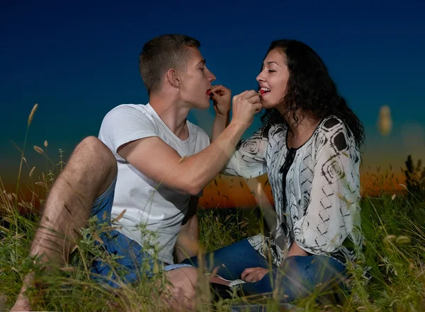 Casal romântico comer bagas vermelhas, sentar-se na grama ao pôr do sol ao ar livre, céu escuro noite, conceito de amor, jovens adultos — Fotografia de Stock