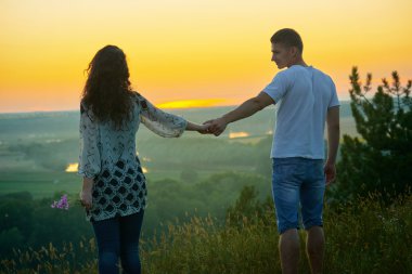 romantik çift ülke açık gün batımında yürüyüş, yabani çiçek buketi ile kız, güzel manzara ve parlak sarı gökyüzü, sevgi hassasiyet kavramı, genç yetişkin insanlar