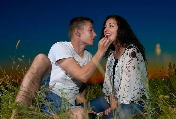 couple eating red raspberry, sit on grass at sunset on outdoor, dark night sky, love concept, romantic young adult people