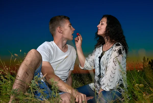 young couple eat red raspberry, sit on grass at dark night sky on outdoor, love concept, romantic young adult people