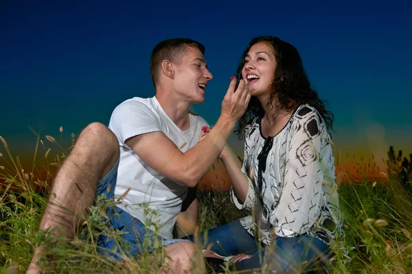 Casal romântico comer framboesa vermelha, sentar-se na grama ao pôr do sol ao ar livre, céu escuro noite, conceito de amor, jovens adultos — Fotografia de Stock