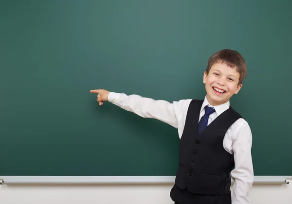 School student boy posing at the clean blackboard, show finger and point at, grimacing and emotions, dressed in a black suit, education concept, studio photo — Stock Photo, Image