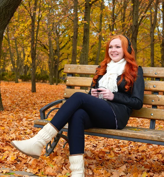 Dívka v podzim sezóny poslouchat hudbu na audio přehrávač se sluchátky, sedí na lavičce v parku, žluté stromy a spadané listí — Stock fotografie