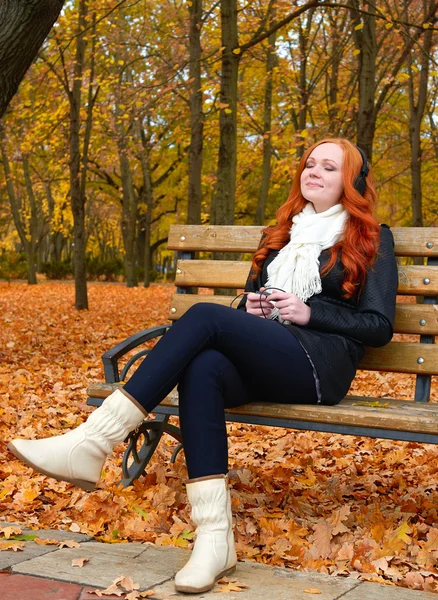 Girl listen music on audio player with headphones, sit on bench in autumn city park, yellow trees and fallen leaves — Stock Photo, Image