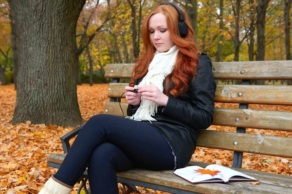 Girl listen music on audio player with headphones, sit on bench in city park, autumn season, yellow trees and fallen leaves — Stock Photo, Image