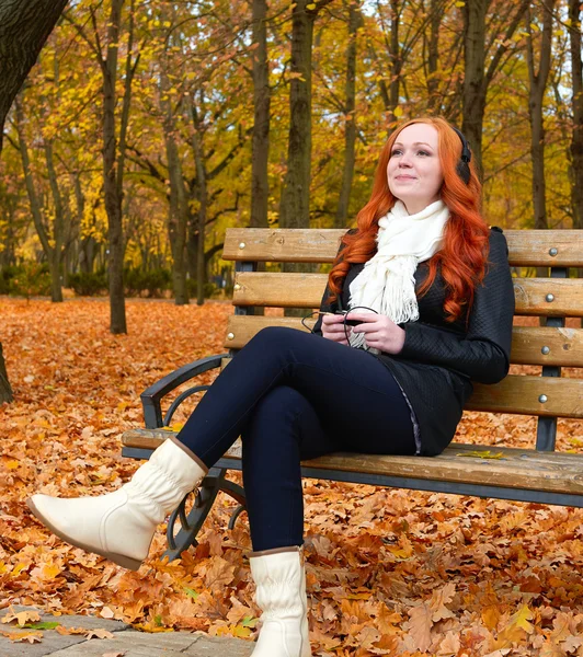 Menina na temporada de outono ouvir música no leitor de áudio com fones de ouvido, sentar-se no banco no parque da cidade, árvores amarelas e folhas caídas — Fotografia de Stock