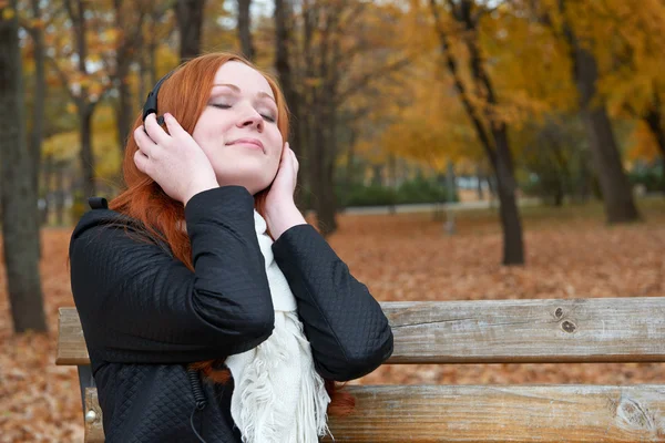 Портрет дівчини, слухати музику на аудіопрогравачі з навушниками, сидіти на лавці в міському парку, осінній сезон, жовті дерева і опале листя — стокове фото