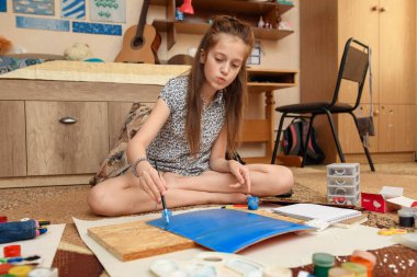 Kız evde resim çiziyor, sanat eseri yapıyor, kağıt, boya ve fırçalardan yaratıcı sanat eserleri yapıyor.