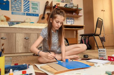 Kız evde resim çiziyor, sanat eseri yapıyor, kağıt, boya ve fırçalardan yaratıcı sanat eserleri yapıyor.