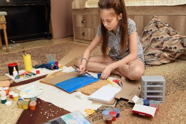 Kız evde resim çiziyor, sanat eseri yapıyor, kağıt, boya ve fırçalardan yaratıcı sanat eserleri yapıyor.